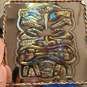 May include: A square metal artwork with a detailed, welded design resembling a stylized human torso. The artwork features a copper-coloured frame with a twisted rope border and a small, arched handle at the top.