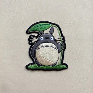 May include: Embroidered patch featuring a gray Totoro character holding a green leaf. The patch has a black border and is set against a beige background. The Totoro has a cream-colored belly and blue eyes.