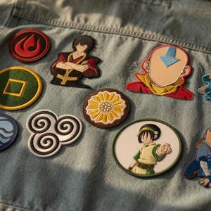 Può includere: Giacca di jeans blu chiaro ornata da varie toppe ricamate colorate. Le toppe presentano personaggi e simboli di una popolare serie animata, tra cui elementi di fuoco, terra, acqua e aria.