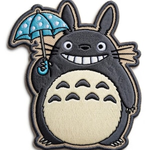 Puede incluir: Parche bordado con un personaje de dibujos animados gris sonriente con ojos grandes y una barriga color crema. El personaje sostiene un paraguas azul con lunares blancos. El parche tiene un borde beige.