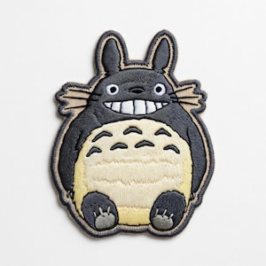 May include: Embroidered patch featuring a smiling, cartoon-style Totoro character. The patch has a gray body, cream-colored belly, and black accents. The design includes small feet, pointed ears, and a detailed outline.