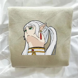 May include: Beige sweatshirt featuring an embroidered anime-style character with white hair, green eyes, and long pointed ears. The character wears a red earring and a gold and red collar. The sweatshirt is folded.