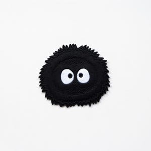 May include: A black, round embroidered patch with a fuzzy texture and two large, white, cartoon-style eyes. The patch has a jagged edge and is set against a white background. Suitable for clothing or accessories.