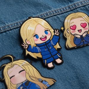 May include: Embroidered patches featuring a cartoon character with blonde hair and blue eyes. The character is wearing a blue jacket and making a peace sign. Other patches show the character with hearts in their eyes and a smiling face.