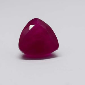 May include: A triangular, deep magenta gemstone with multiple facets. The polished surface of the gem reflects light, creating highlights and shadows. The gem is set against a plain white background. The gem is likely a ruby.
