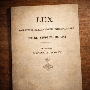 Op de afbeelding: Een vintage boekomslag met de titel "LUX" in grote, vette letters. De omslag bevat Italiaanse tekst, waaronder "BOLLETTINO DELL'ACCADEMIA INTERNAZIONALE" en "PER GLI STUDI PSICOLOGICI." De naam van de directeur, "GIOVANNI HOFFMANN", is ook zichtbaar.