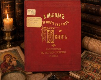 Puede incluir: Un libro de tapa dura rojo con letras cirílicas doradas en la portada, que presenta una pequeña etiqueta de papel rectangular con texto escrito a mano. El libro está colocado en posición vertical, mostrando una inicial decorativa y datos.