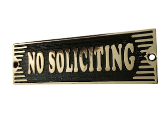 May include: A rectangular sign with the words "NO SOLICITING" in gold lettering. The sign has a black background with gold trim and two holes for mounting. The sign is designed to be mounted on a door or wall.