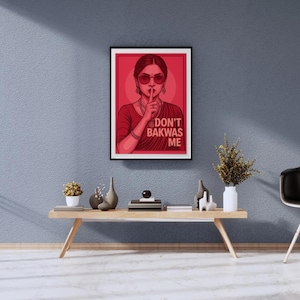 May include: Framed red and black art print featuring a woman in sunglasses with a finger to her lips. The text "DON'T BAKWAS ME" is displayed on the print. The artwork is displayed on a blue wall above a wooden table.