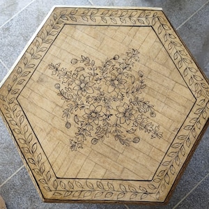 Puede incluir: Mesa hexagonal de madera con acabado marrón claro. La mesa presenta un diseño floral en el centro y un borde de hojas. La mesa mide aproximadamente 61 cm de ancho.