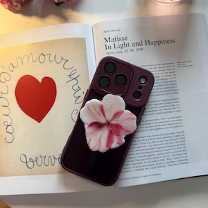 Può includere: Una custodia per telefono bordeaux con un supporto a forma di fiore bianco e rosa, appoggiata su un libro aperto. Il titolo del libro è "Matisse In Light and Happiness". È visibile anche una stampa incorniciata con un cuore rosso e la parola "amour".