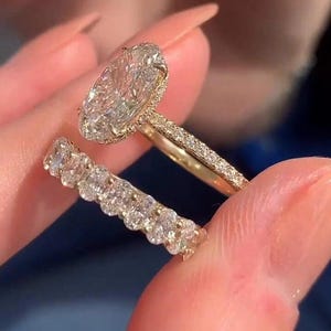 Può includere: Un anello di fidanzamento in oro con un grande diamante taglio ovale e una fascia tempestata di diamanti. È visibile anche una fede nuziale con diamanti abbinata. Gli anelli sono tenuti su uno sfondo sfocato.