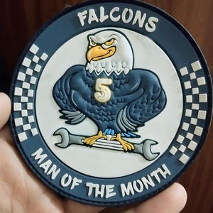 May include: A round patch with a blue border and the word "FALCONS" in white. The patch features an eagle with a "5" on its chest, standing on a wrench. The text "MAN OF THE MONTH" is also on the patch.