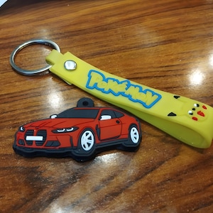 Könnte beinhalten: Ein gelbes Silikon-Schlüsselanhängerband mit dem blauen Pokémon-Logo und einem kleinen Pikachu-Gesicht, befestigt an einem silbernen Metallring. Daneben liegt ein roter Gummi-Anhänger in Form eines Sportwagens mit weißen Rädern.