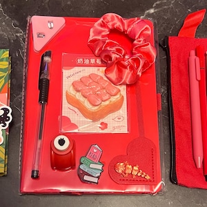 Peut inclure: Ensemble de fournitures de bureau rouges et roses. Comprend un agenda avec des autocollants, un cahier rouge, un chouchou, des stylos, une trousse et des gommes. L'agenda porte l'inscription "2026-27".