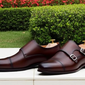 May include: A pair of brown leather dress shoes with a cap toe design. One shoe features a double monk strap with silver buckles, while the other is a slip-on style. The shoes are displayed on a white surface.
