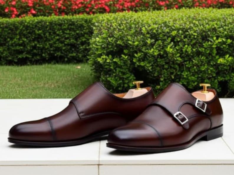 May include: A pair of brown leather dress shoes with a cap toe design. One shoe features a double monk strap with silver buckles, while the other is a slip-on style. The shoes are displayed on a white surface.
