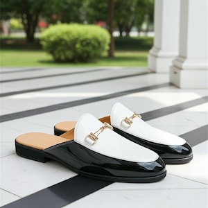 May include: Pair of black and white leather slip-on shoes with gold-colored metal accents. The shoes have a glossy finish and a classic design, suitable for various occasions. The soles are black, and the inner sole is tan.