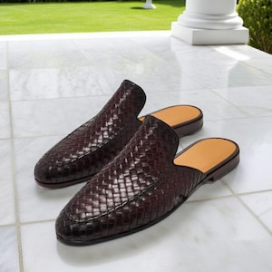 May include: A pair of dark burgundy woven leather slip-on shoes. The shoes have a rounded toe and a low heel. The interior is a light tan color. The shoes are placed on a white marble surface.