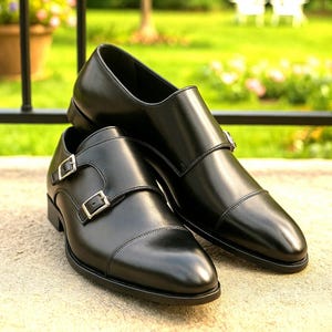May include: A pair of black leather monk strap shoes with silver buckles. The shoes have a polished finish and a classic design, suitable for formal or business wear. The shoes are displayed on a concrete surface with a blurred green background.