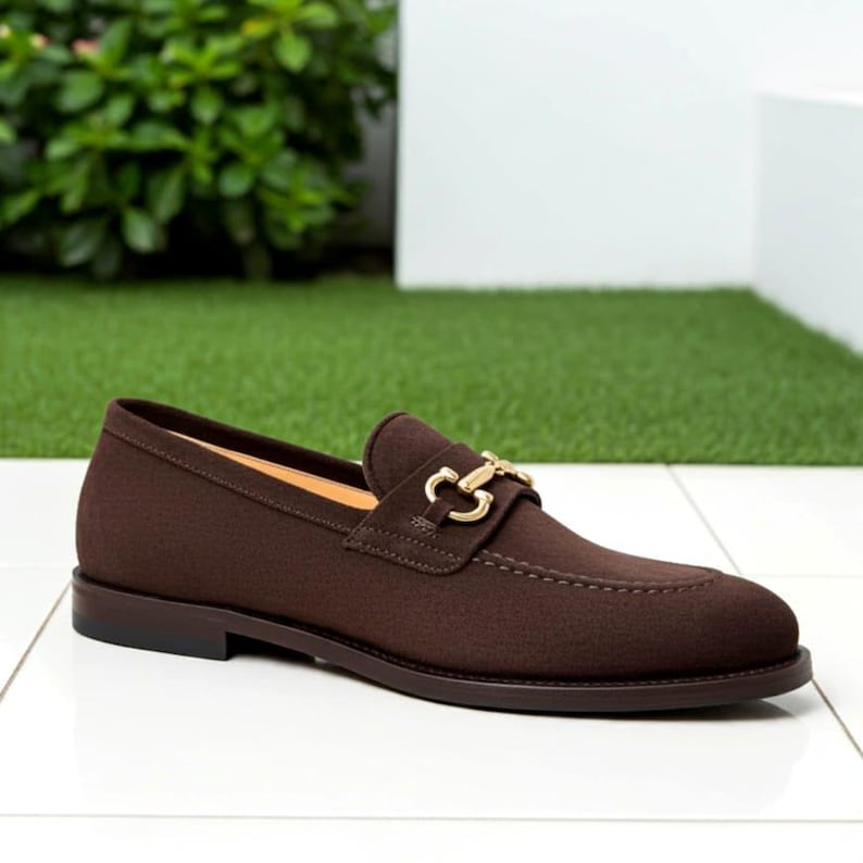 May include: A brown suede loafer with a gold-colored buckle. The shoe has a rounded toe and a dark brown sole. The loafer is displayed on a white tile surface with a green background.