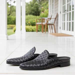 May include: A pair of black woven leather slip-on shoes. The shoes have a pointed toe and a low heel. The woven pattern covers the entire upper of the shoe. The shoes are displayed on a white tiled surface.