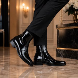 May include: A pair of black patent leather ankle boots with a high-gloss finish, featuring a side zipper closure, a rounded toe, and a low stacked heel. The boots are worn with black trousers, showcasing a sleek, polished, and reflective surface.