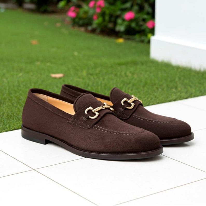 May include: A pair of brown suede loafers with gold-colored metal accents. The shoes have a classic design with a rounded toe and a low heel. The loafers are displayed on a white tiled surface.