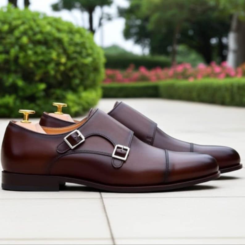 May include: A pair of brown leather double monk strap shoes with silver buckles. The shoes have a classic design with a cap toe and are displayed on a light-colored surface. The background features greenery.