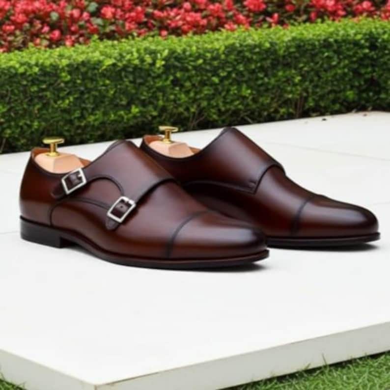 May include: A pair of brown leather double monk strap shoes with silver buckles. The shoes are displayed on a white surface with a green and red floral background. The shoes have wooden shoe trees inside.
