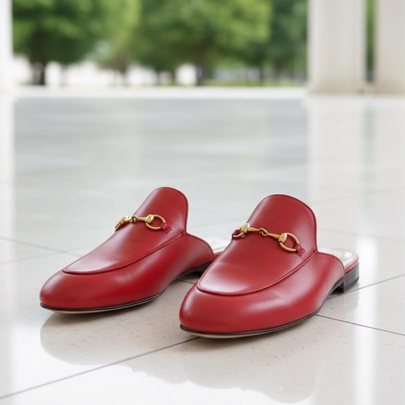 Op de afbeelding: Een paar rode leren instappers zonder veters met gouden metalen bitdetail. De schoenen hebben een ronde neus en een lage hak. De loafers worden gepresenteerd op een wit betegeld oppervlak.
