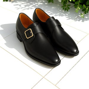 May include: A pair of black leather monk strap shoes with a gold-colored buckle. The shoes have a sleek, polished appearance and a classic design. The interior lining is a light tan color.