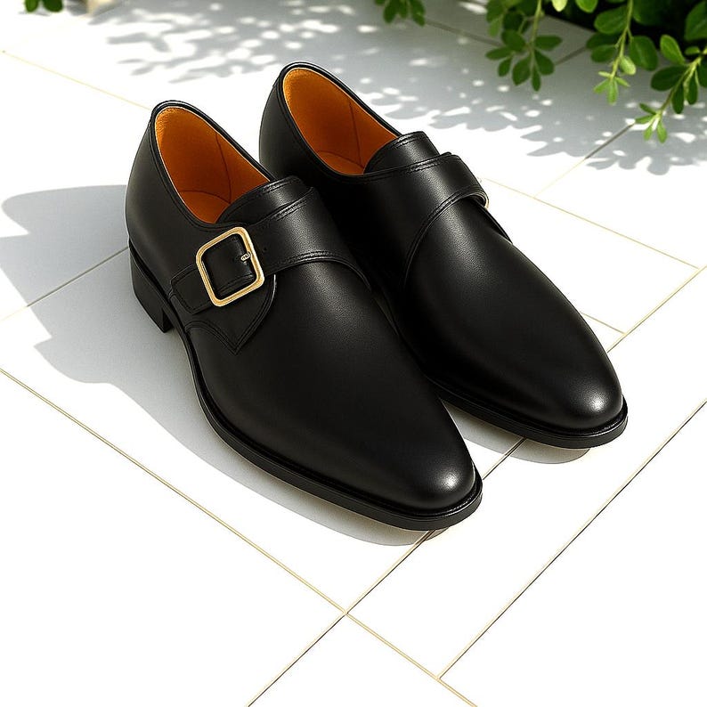 May include: A pair of black leather monk strap shoes with a gold-colored buckle. The shoes have a sleek, polished appearance and a classic design. The interior lining is a light tan color.