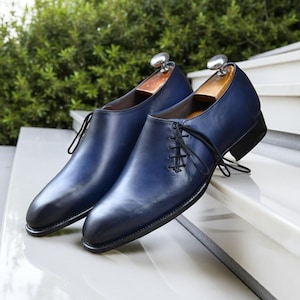 K&ouml;nnte beinhalten: Ein Paar marineblaue Lederschuhe mit schwarzen Sohlen und Schn&uuml;rsenkeln. Die Schuhe werden auf einer wei&szlig;en Oberfl&auml;che mit gr&uuml;nem Hintergrund pr&auml;sentiert. Die Schuhe haben Schuhspanner aus Holz.