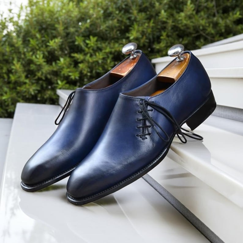 K&ouml;nnte beinhalten: Ein Paar marineblaue Lederschuhe mit schwarzen Sohlen und Schn&uuml;rsenkeln. Die Schuhe werden auf einer wei&szlig;en Oberfl&auml;che mit gr&uuml;nem Hintergrund pr&auml;sentiert. Die Schuhe haben Schuhspanner aus Holz.