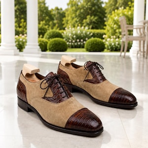 May include: A pair of oxford shoes featuring a combination of tan suede and dark brown crocodile-embossed leather. The shoes have a cap toe design, dark brown laces, and light-coloured wooden shoe trees inserted into the openings for shape.