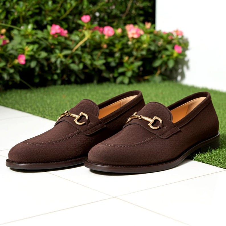 May include: A pair of brown suede loafers with gold-colored metal accents. The shoes have a classic design with a rounded toe and a slip-on style. The loafers are displayed on a white tiled surface with a green background.