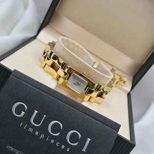 Puede incluir: Un reloj Gucci dorado con esfera cuadrada y pulsera de eslabones dorados. Se muestra en su caja negra original, con el logotipo de Gucci y la palabra "timepieces" en una tarjeta.