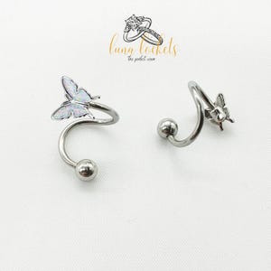 May include: Two silver-colored spiral earrings, each with a small ball at the base and a butterfly design at the top. The butterfly has a pearlescent, iridescent appearance. The earrings are displayed on a white background. The brand name "Luna Lockels" is visible.