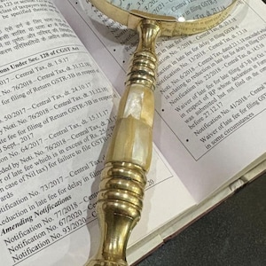 May include: A vintage-style magnifying glass with a brass handle and a clear lens, resting on an open book with printed text. The handle features a decorative design with a mother-of-pearl inlay.