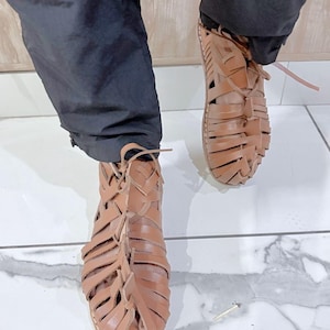 Puede incluir: Sandalias de cuero marrón con un diseño tejido. Las sandalias tienen una suela plana y se atan con cordones en la parte delantera. La imagen muestra las sandalias desde un ángulo bajo, con las piernas del usuario visibles por encima.