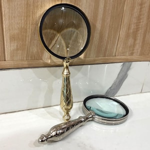 May include: Two magnifying glasses with ornate handles. One has a gold-colored handle and a black rim around the lens. The other has a silver-colored handle and a black rim around the lens. Both are on a white surface.