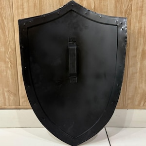 Puede incluir: Un escudo negro con una parte superior curvada y un asa en la parte posterior. El escudo tiene una superficie ligeramente texturizada y est&aacute; bordeado con remaches. El escudo es de metal y probablemente sea para fines decorativos o hist&oacute;ricos.