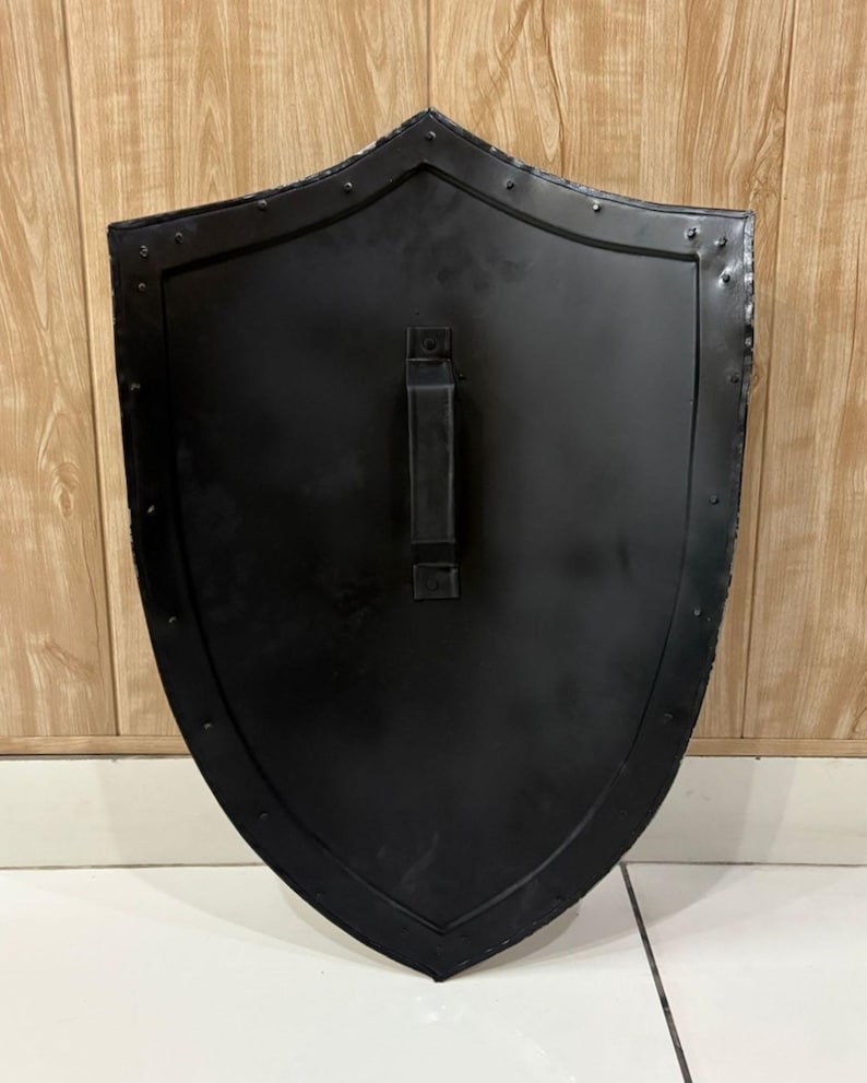 Puede incluir: Un escudo negro con una parte superior curvada y un asa en la parte posterior. El escudo tiene una superficie ligeramente texturizada y est&aacute; bordeado con remaches. El escudo es de metal y probablemente sea para fines decorativos o hist&oacute;ricos.