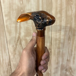 May include: A wooden walking stick featuring a rabbit head handle. The rabbit head is bronze and black, with a long ear. The shaft is light brown with a twisted design. The cane is held by a person.