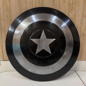 May include: A black and silver shield featuring a central silver star. The shield has a circular design with black and silver concentric rings. The star is positioned on a black background.