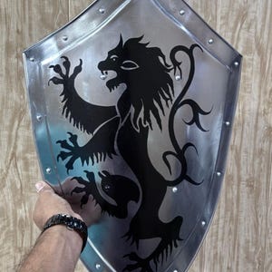 Pu&ograve; includere: Uno scudo color argento con un emblema di leone nero. Lo scudo ha un bordo rivettato e una forma curva e appuntita. Il leone &egrave; raffigurato in stile araldico, con un'espressione feroce.
