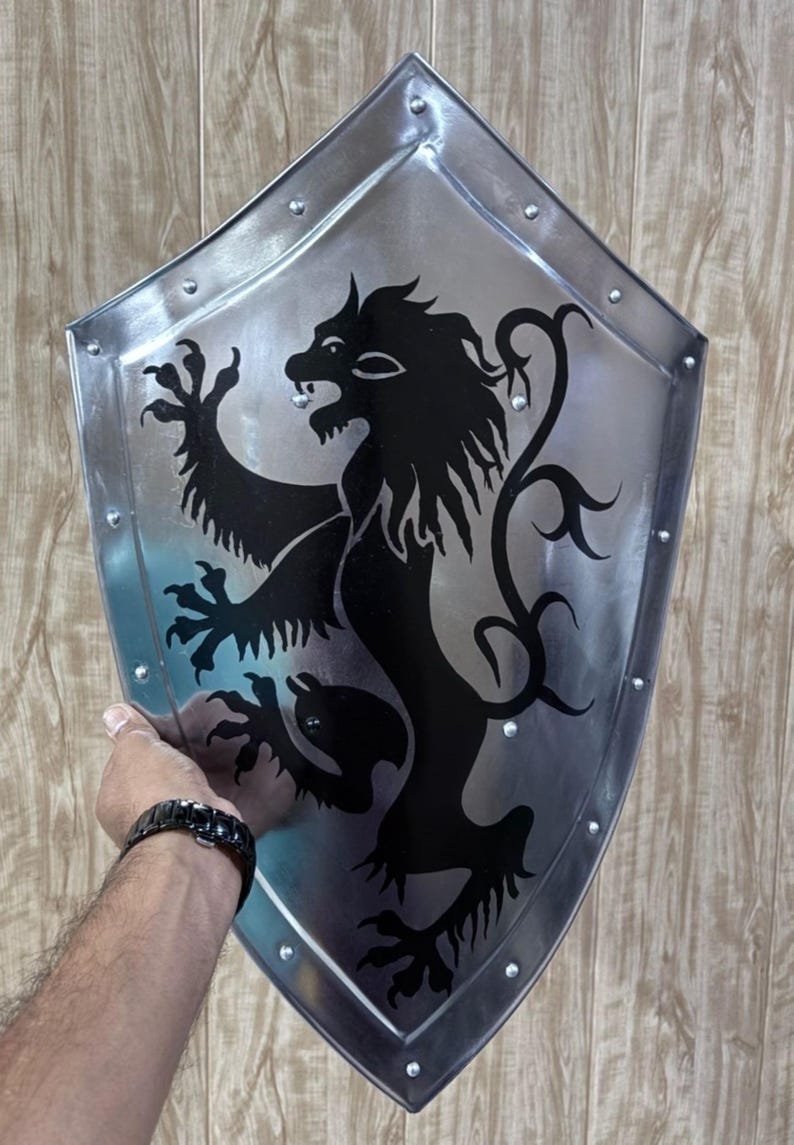 Pu&ograve; includere: Uno scudo color argento con un emblema di leone nero. Lo scudo ha un bordo rivettato e una forma curva e appuntita. Il leone &egrave; raffigurato in stile araldico, con un'espressione feroce.