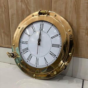 Puede incluir: Un reloj de pared circular con un marco de latón pulido en forma de ojo de buey, con remaches decorativos y dos pestillos laterales. La esfera es blanca con números romanos negros y manecillas finas negras, incluyendo un segundero de color rojo.