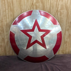May include: A circular shield with a red and silver design. The shield has a large red star in the centre, set against a silver background and a red outer ring. The shield is a prop or collectible.